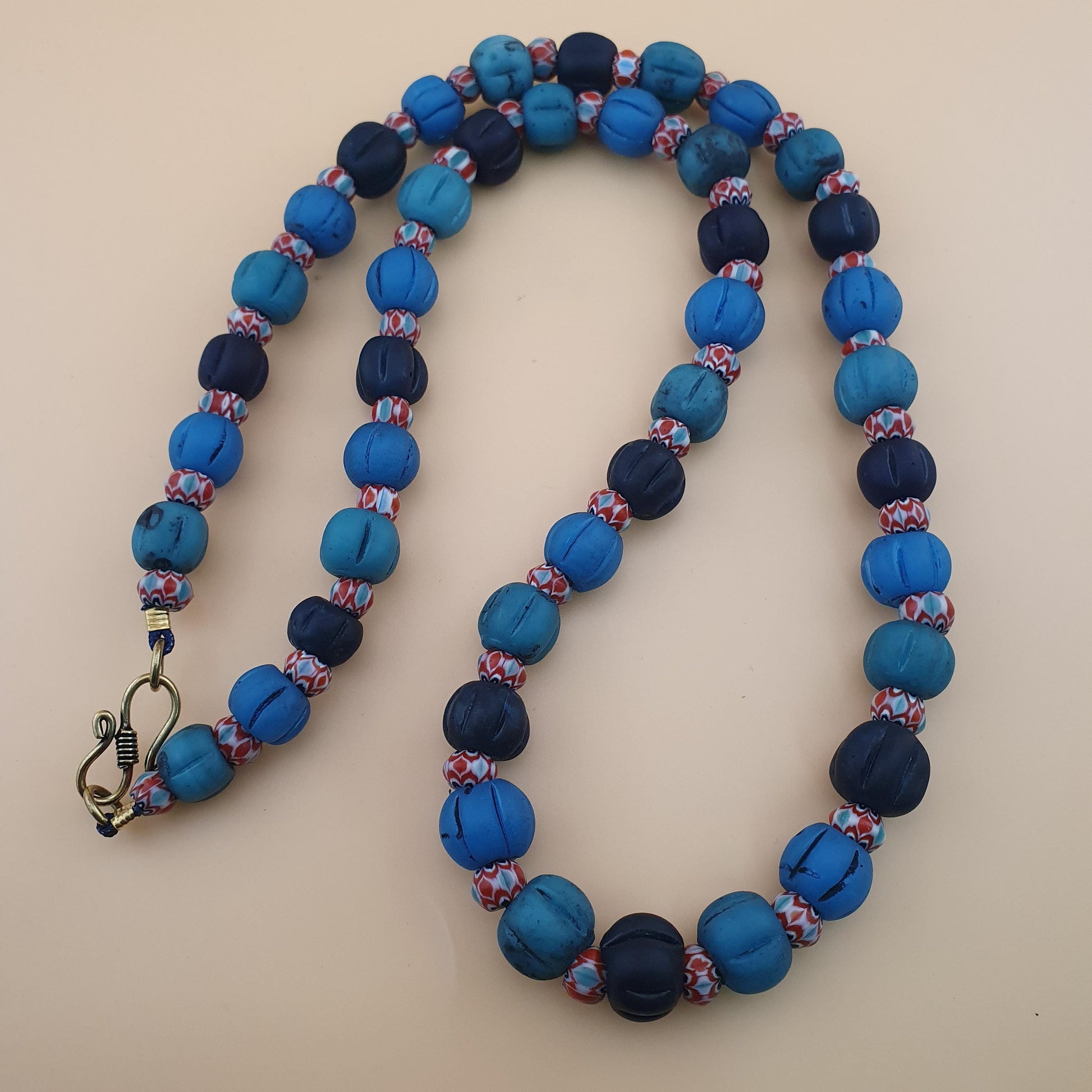 A beaded necklace with alternating large textured blue and navy beads, separated by smaller marbled blue, red, and white beads, featuring a silver-toned lobster clasp on a light beige background.