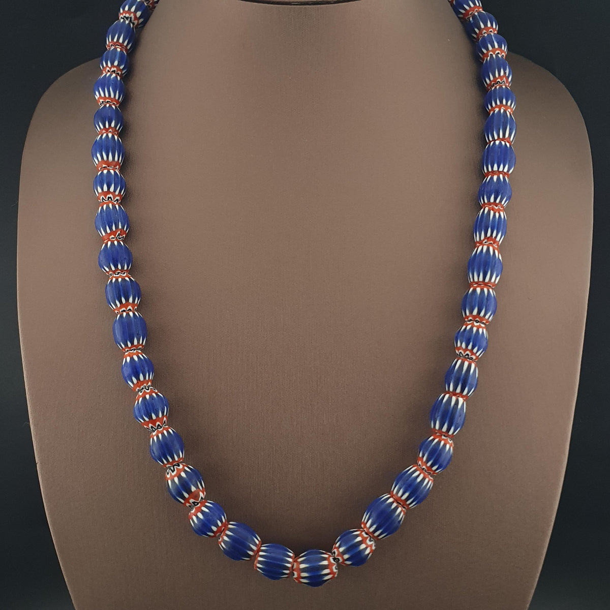 A beaded Chevron necklace displayed on a brown mannequin bust. The necklace features glossy blue oval beads with white spiral patterns and thin orange-red borders, 