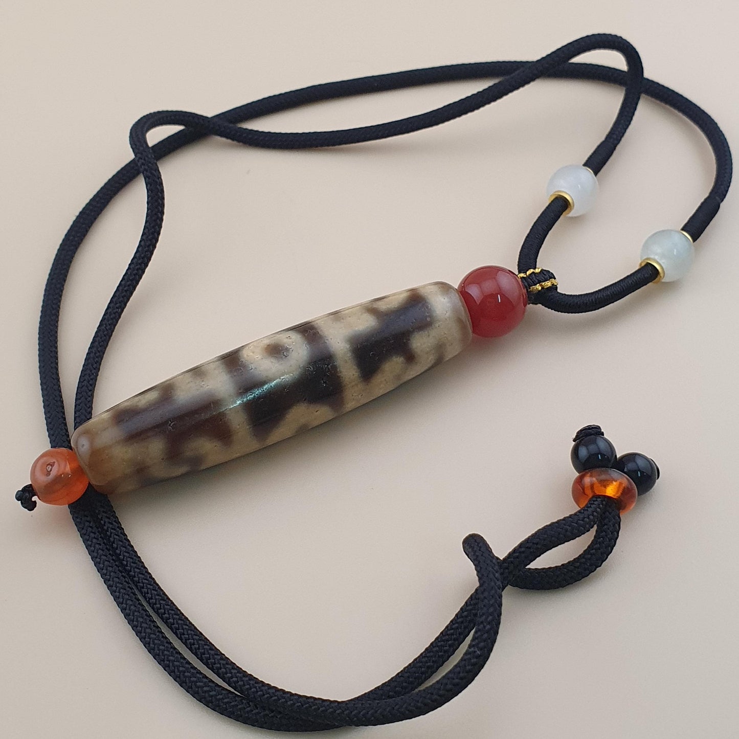 A beaded necklace featuring a central tiger's eye bead with brown and cream bands, flanked by a red bead and light blue beads on a black cord, set against a solid beige background.