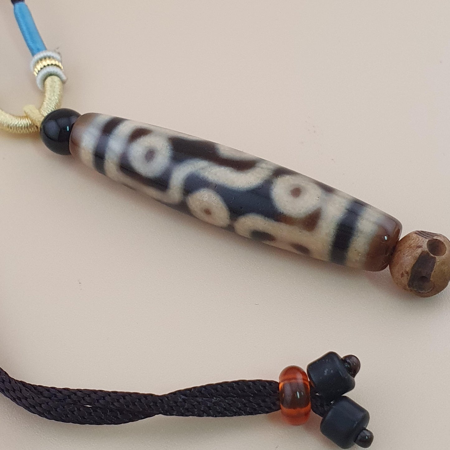 A traditional Tibetan dawa bead pendant with a black and cream pattern, threaded on a black cord with a blue accent cord and silver spacer, displayed on a beige background.