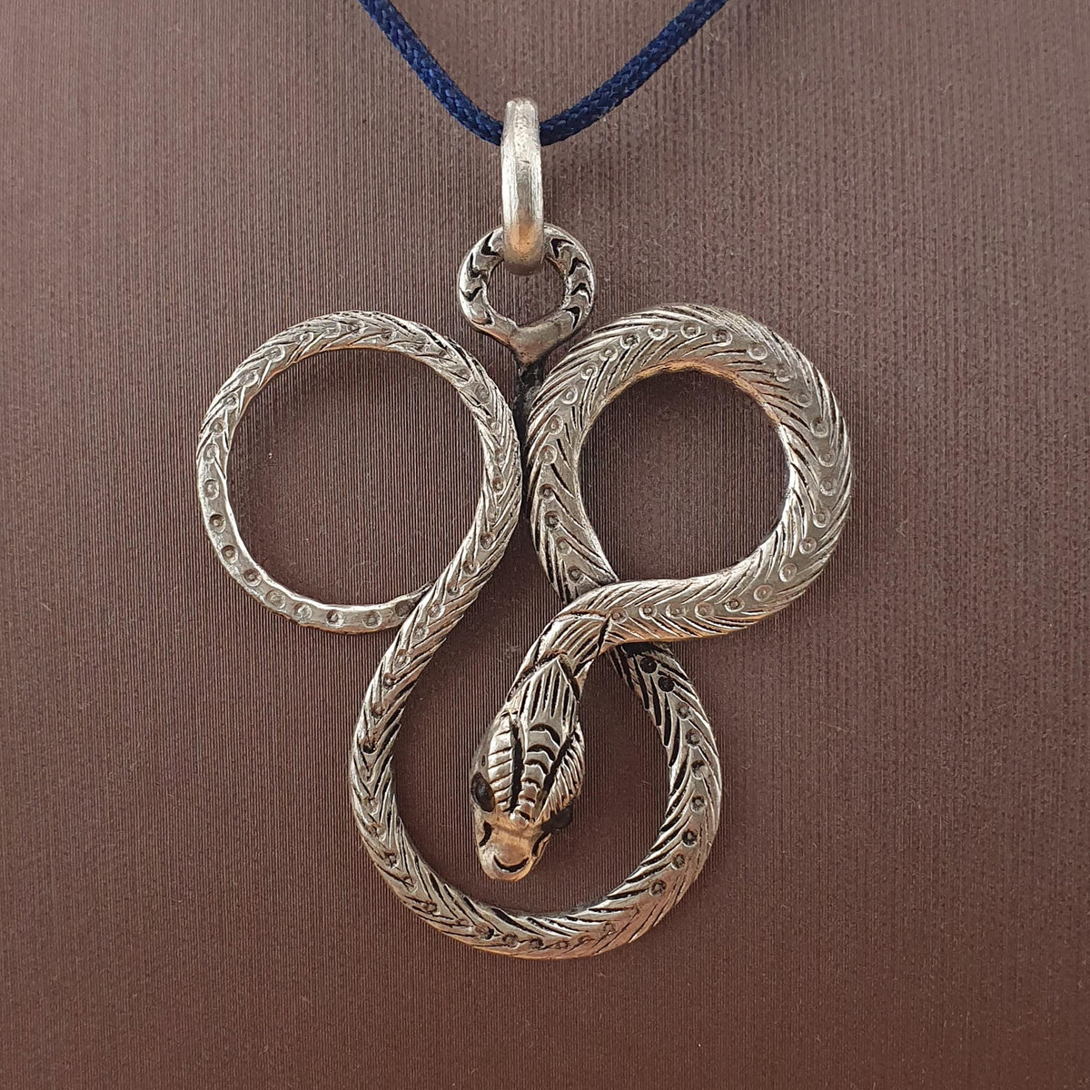A Brass silver pendant necklace featuring a coiled snake design with an engraved head and a dark blue cord, displayed on a textured brown background.
