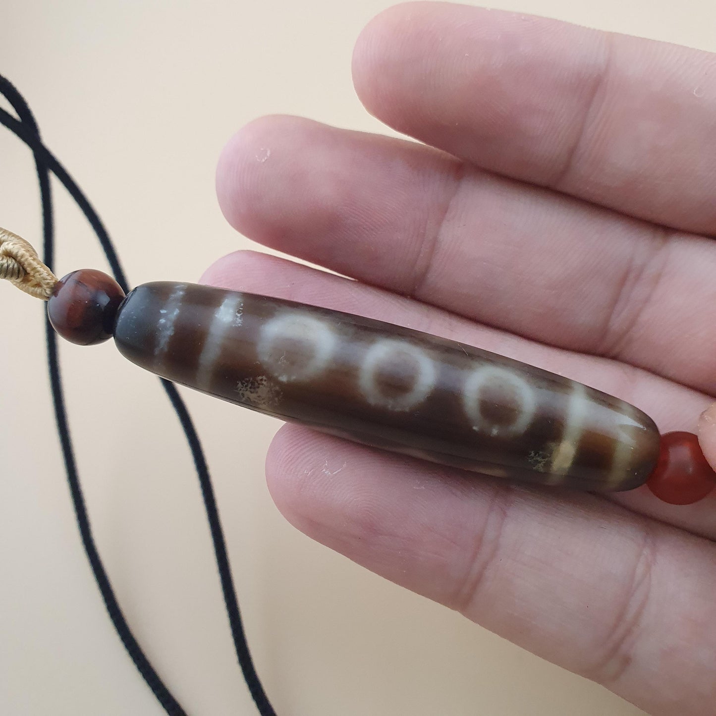 A close-up of a hand holding a brown agate bead pendant with white markings and a black cord, against a plain beige background.